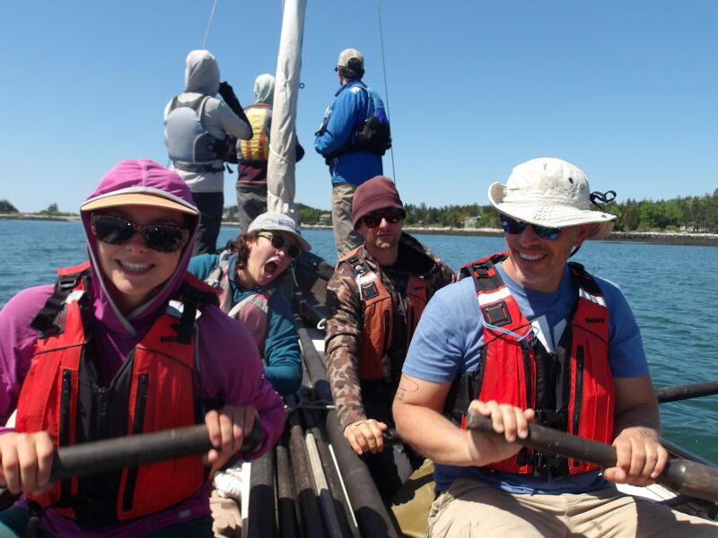 The image shows a group of people on a boat, likely rowing. They are wearing life jackets and sunglasses, enjoying a sunny day on the water. The background includes the sky and some trees. The people seem happy and engaged in the activity.